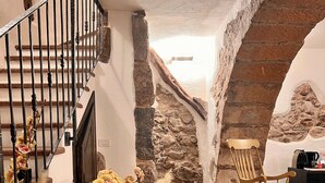 Ein Steinkunstwerk führt in einen Raum mit einem Holzschaukelstuhl, einer Topfpflanze und einer Treppe mit Metallgeländer.