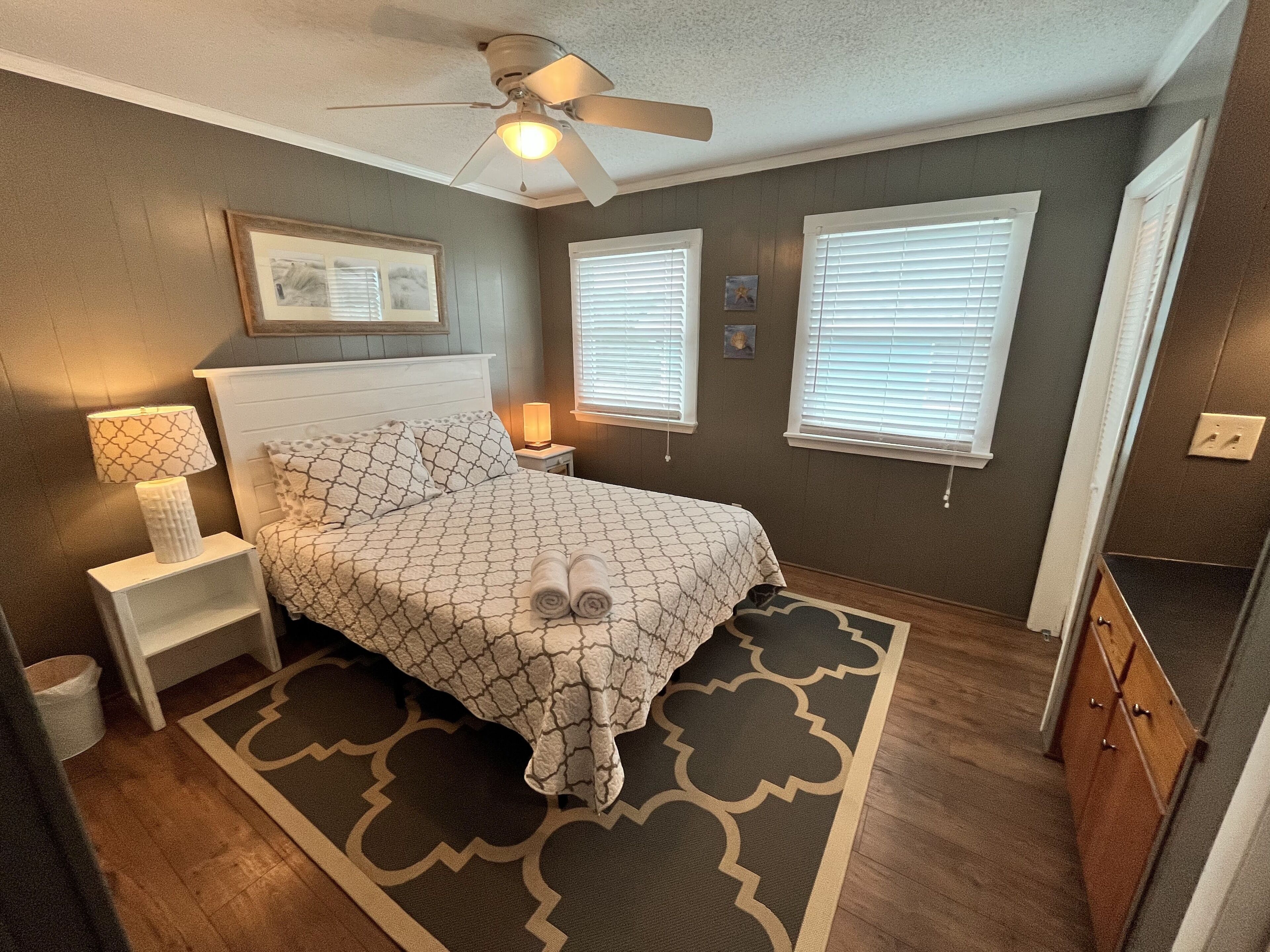A bedroom with a bed, nightstands, a ceiling fan, and two windows with blinds.