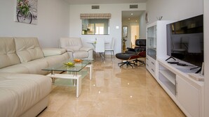 Un salon moderne avec un canapé beige, une table basse en verre, un coin repas avec des chaises blanches et un téléviseur posé sur un meuble.