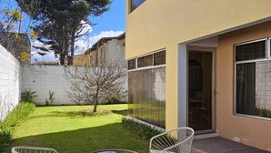 Ett modernt hus med en gul fasad, en liten trädgård med en gräsmatta och två vita metallstolar runt ett glasbord.