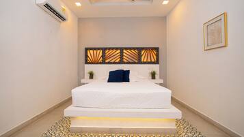 Un dormitorio con cama, cabecera con paneles de luz, una planta pequeña y un cuadro en la pared.