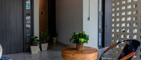 Una habitación con una mesa redonda de madera, una silla tejida azul y una silla negra con detalles de madera. Hay una planta en maceta sobre la mesa y un panel decorativo en la pared con cuadrados recortados.