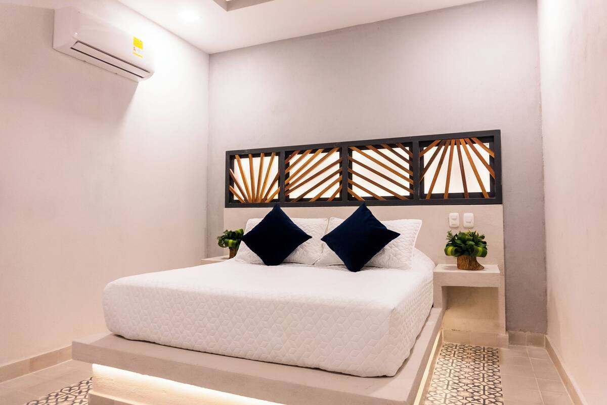 Une chambre moderne avec un grand lit, une tête de lit décorative, des tables de chevet ornées de plantes et un appareil de climatisation.