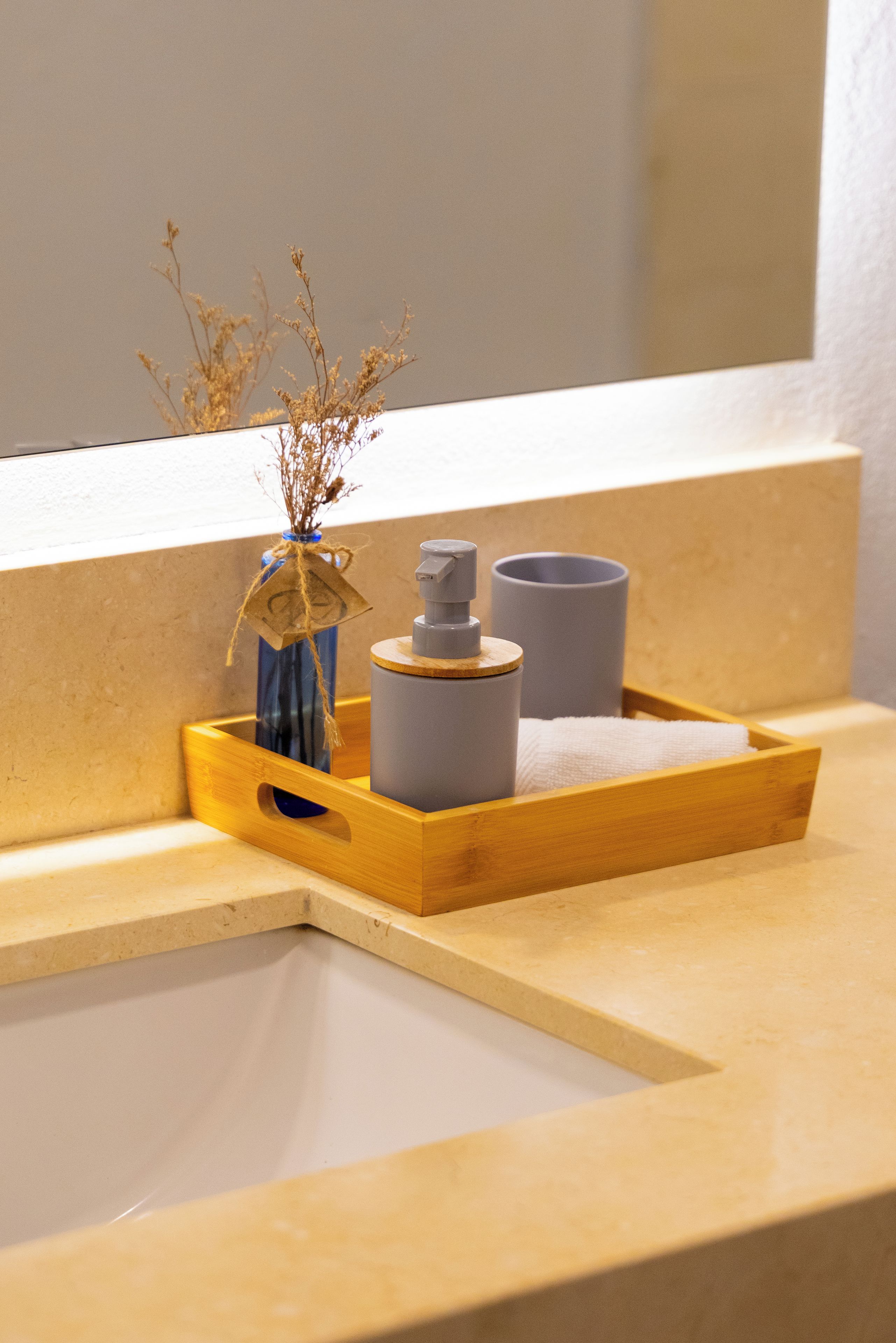 Ein Badezimmer-Waschtisch mit einer Holztablett, auf dem eine Vase mit getrockneten Blumen, ein Seifenspender und eine Tasse stehen.