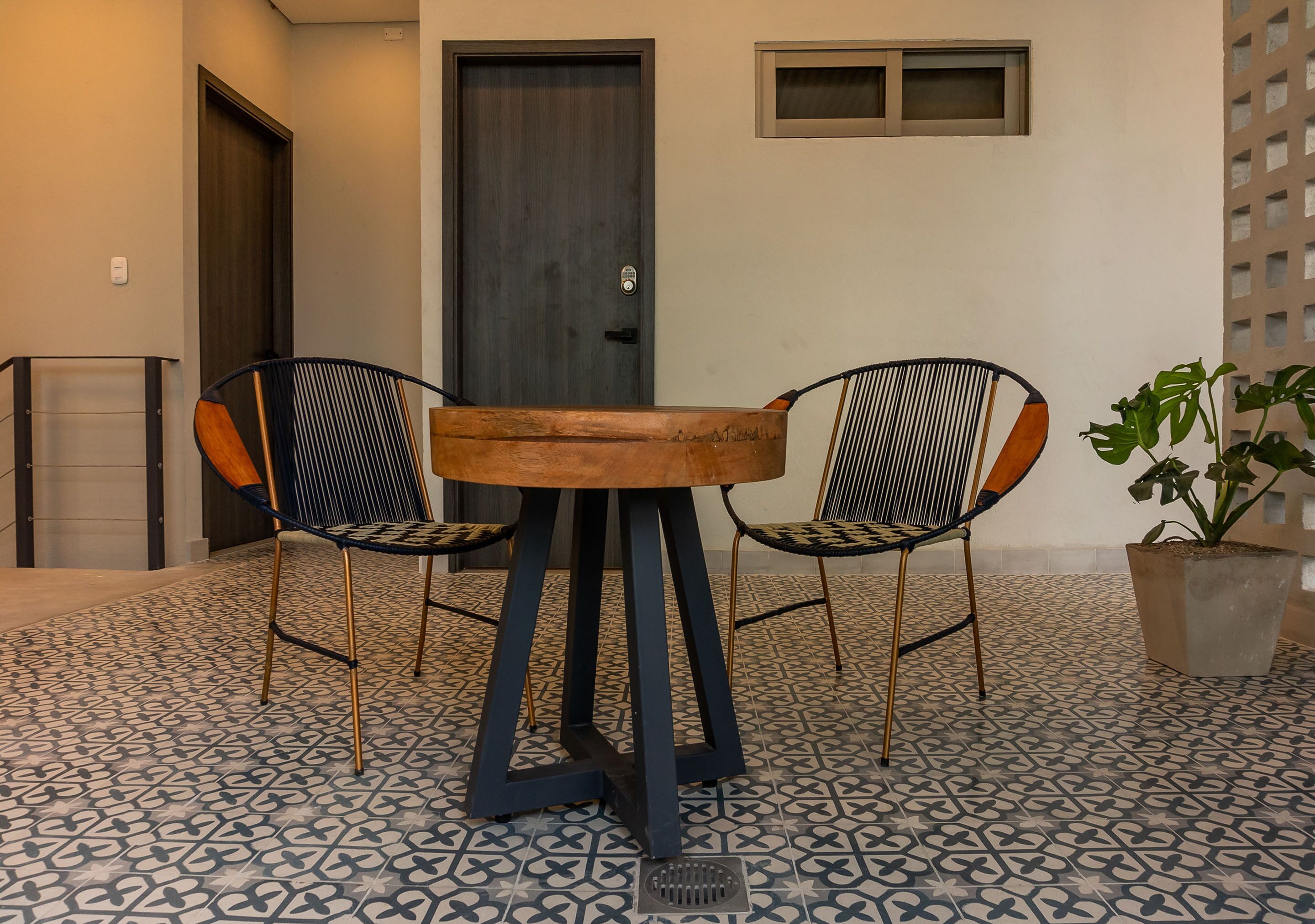Une pièce avec une table ronde en bois et deux chaises à structure métallique et à coussins à motifs. Une plante en pot se trouve sur le sol.