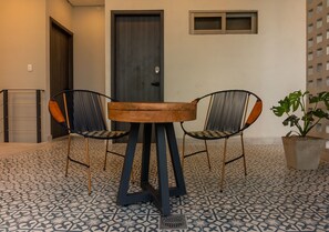 Una habitación con una mesa redonda de madera y dos sillas con estructura metálica y cojines estampados. Hay una planta en maceta en el suelo.