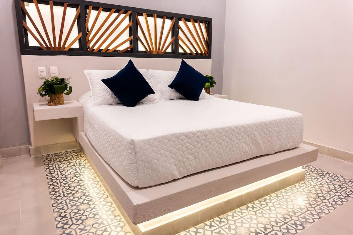 Une chambre à coucher moderne avec un lit blanc, deux oreillers bleu foncé, une table de chevet avec une plante et une tête de lit décorative.
