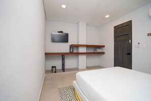 Ein Schlafzimmer mit einem Bett, einem an der Wand montierten Fernseher, einem Holzregal, einer Tür und einem Mülleimer.