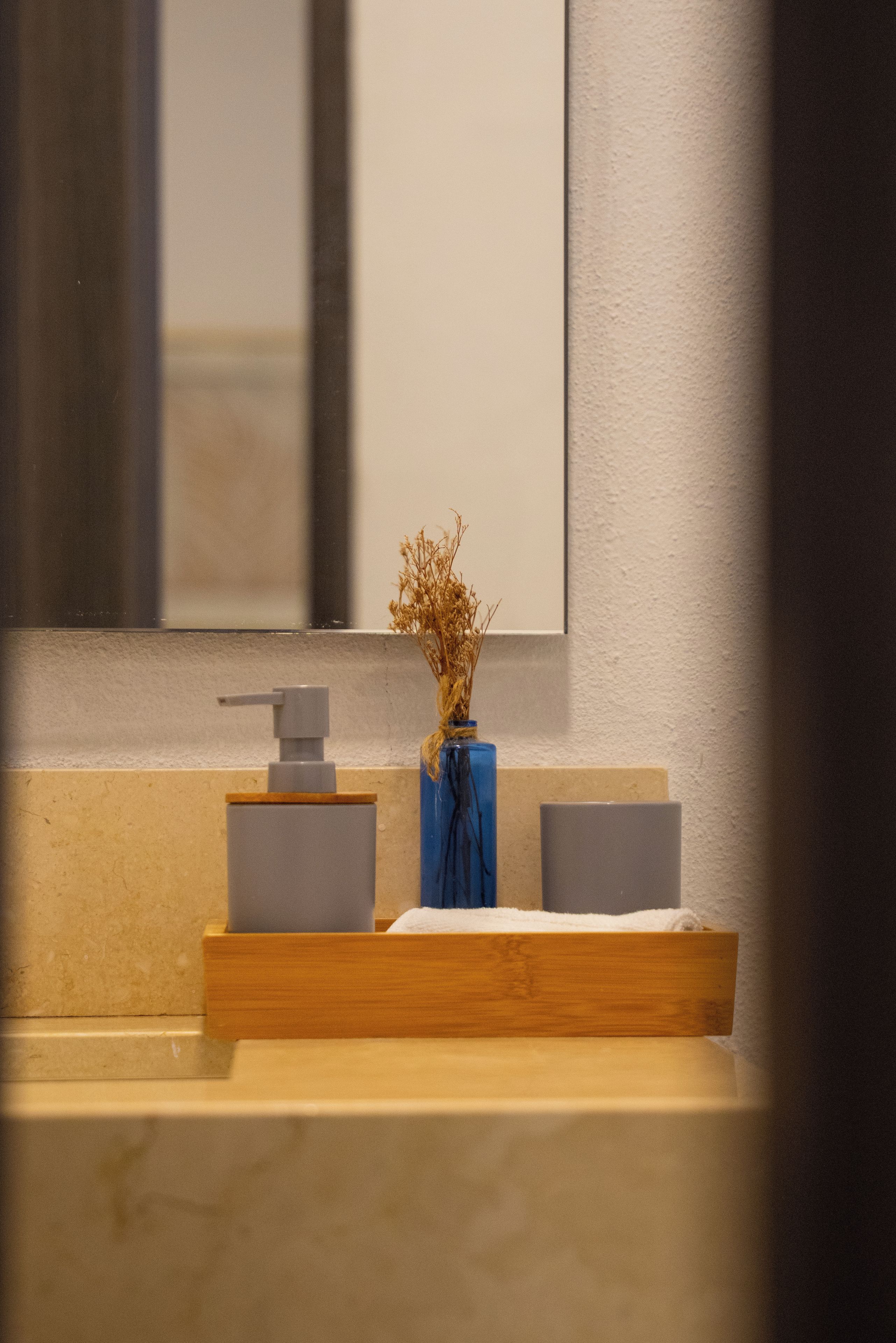 Un baño con espejo, dispensador de jabón y un florero con plantas secas.