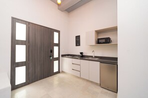 A modern kitchen with a dark wood door, white cabinets, a stainless steel sink, a microwave, and a coffee maker. - The Garden Suites (Santa Marta)