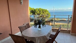 Una mesa redonda con mantel blanco y sillas de madera, un jarrón con flores, y vista al mar y a los árboles.