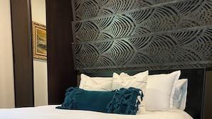 A neatly made bed with white bedding and teal throw pillows, a dark headboard with a geometric pattern, and a partially visible door with a framed picture.