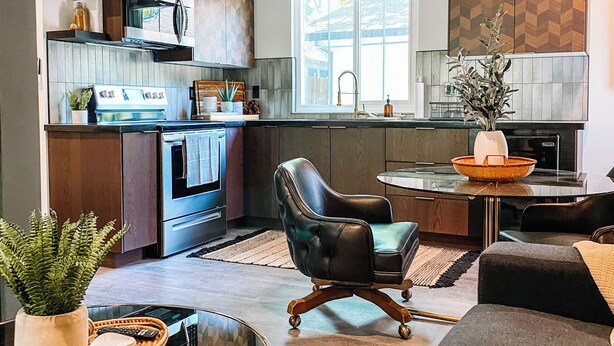 A modern kitchen with dark wood cabinets, a black countertop, and a built-in oven. There's a dining area with a round table, a black leather chair, and a woven rug.