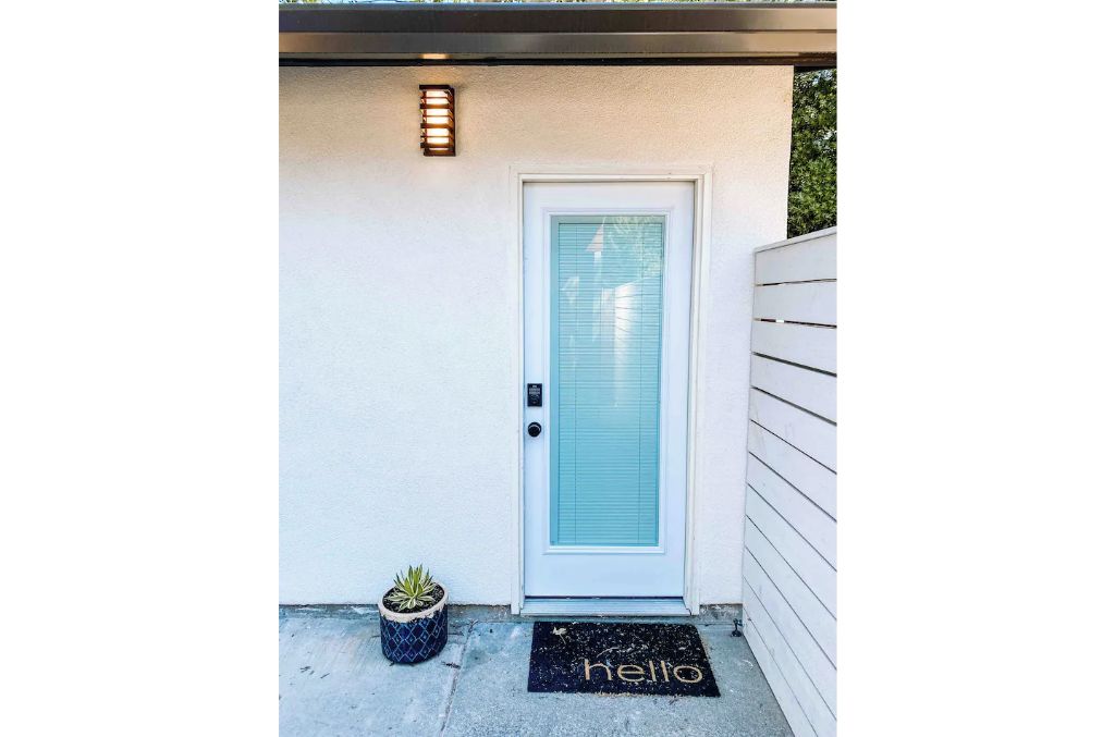 A white door with a frosted glass panel, a small potted plant, and a 'hello' mat on the ground.