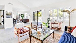 Een ruime woonkamer met een glazen salontafel, houten stoelen, een blauwe bank en grote ramen met zonwering.