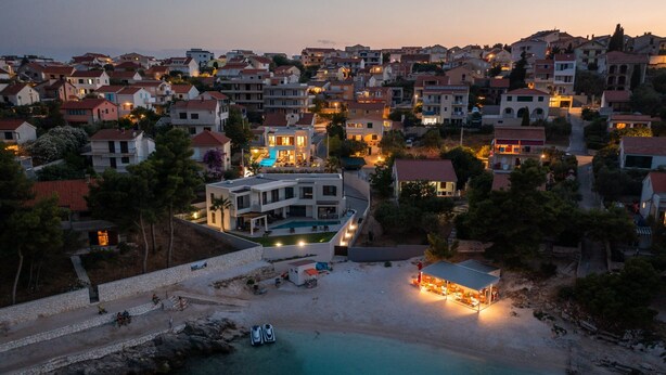 A coastal town at dusk with a prominent modern house featuring a swimming pool, a beach with umbrellas, and a small dock with boats. - Luxury Villa The North Star with Pool (Okrug Gornji)
