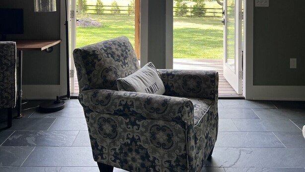A room with a high ceiling, a patterned armchair, a small table, and a view through glass doors to a green outdoor area.