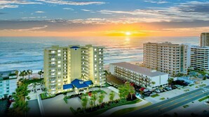 Un complejo hotelero frente al mar con varias torres altas, un estacionamiento y vistas al océano al atardecer.