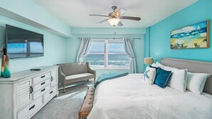 Un dormitorio con cama, una silla, cómoda, ventilador de techo, televisión y ventana con vista al océano.