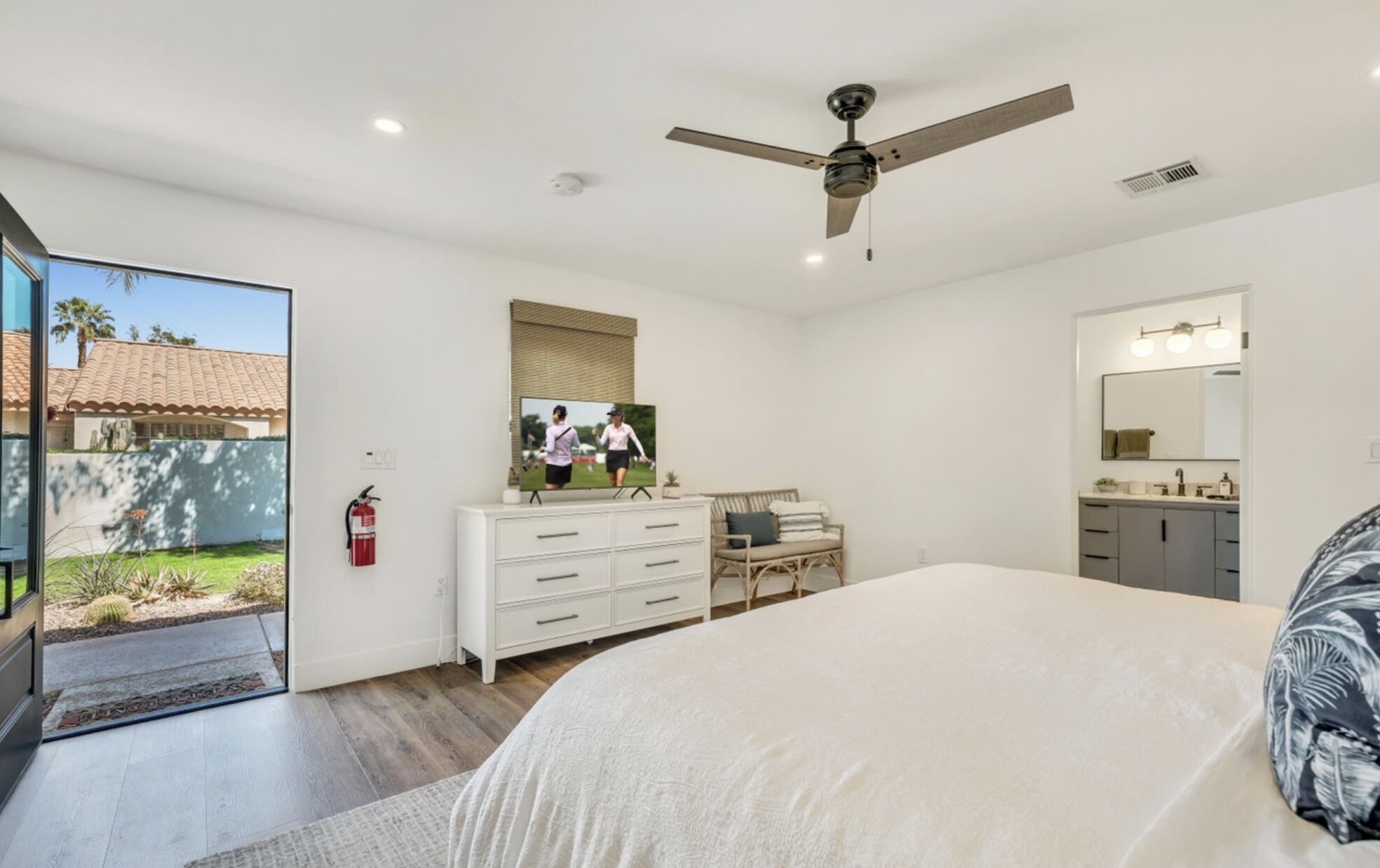 A bedroom with a bed, a dresser, a vanity, a ceiling fan, and a large window overlooking a garden.