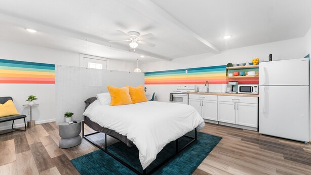Une chambre moderne avec un lit, une table de chevet et une petite plante. À côté se trouve une kitchenette équipée d’un four à micro-ondes, d’un grille-pain et d’un réfrigérateur.
