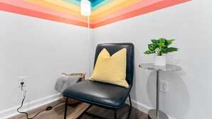Une pièce d’angle avec un fauteuil en cuir noir, un coussin jaune, une petite plante en pot et un plafond blanc orné de rayures arc-en-ciel.