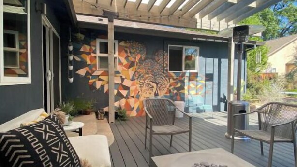 An outdoor patio with a wooden deck, a sofa with patterned cushions, a stone fireplace, and a colorful abstract mural on the wall. - West Park House | Relaxing Park Life (Chico)