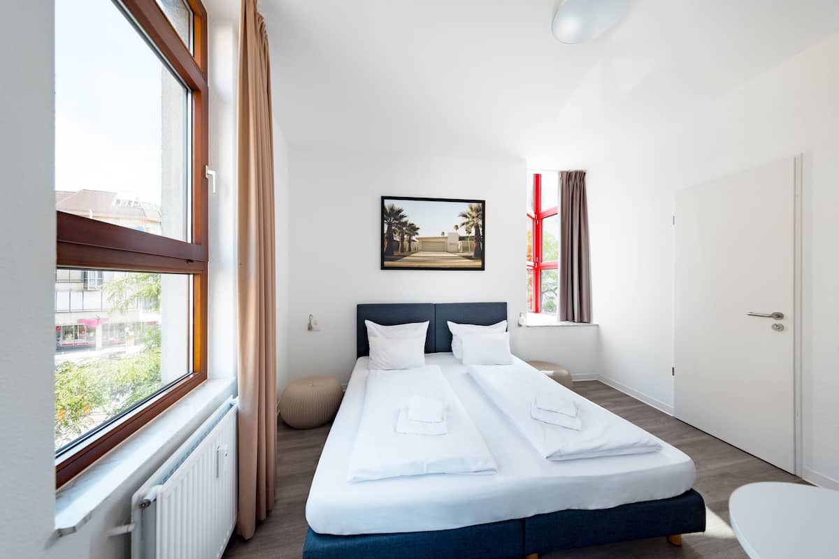 A modern hotel room with a large bed, a window with curtains, a framed picture on the wall, and a door.