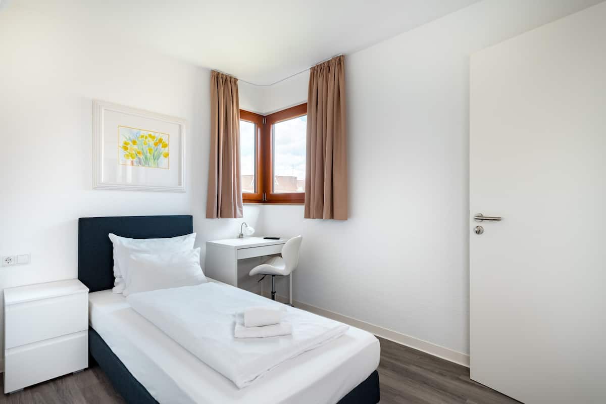 A hotel room with a bed, a desk, a chair, a nightstand, and a door.