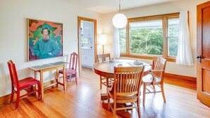 A dining area with a round table, wooden chairs, a painting on the wall, and a window with curtains.