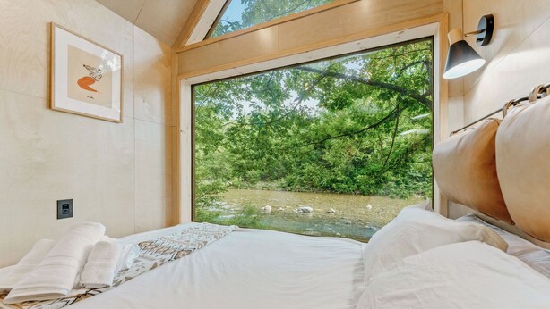 Camera da letto con un'ampia finestra vista su una zona boschiva e un ruscello.