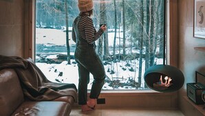 Una persona in piedi vicino a una finestra con vista su una foresta innevata, un divano in pelle e un camino.