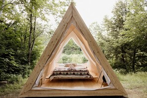 Una tienda de madera triangular con una cama en su interior, rodeada de árboles.