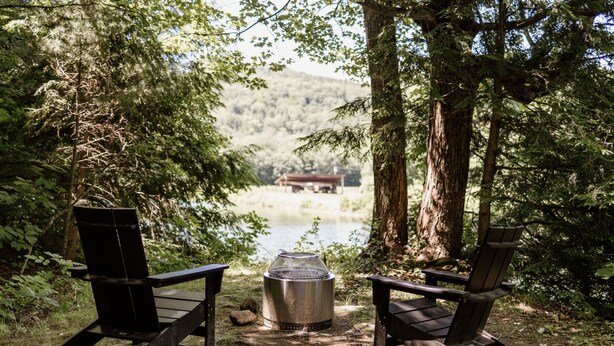 Two Adirondack chairs facing a campfire with a metal pot in the center, surrounded by trees and a lake view.