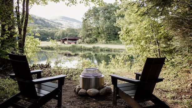 Två Adirondack-stolar placerade framför en lägereld, med en metallgrill i mitten, omgiven av stenar. I bakgrunden syns en sjö och skogbeklädda kullar.