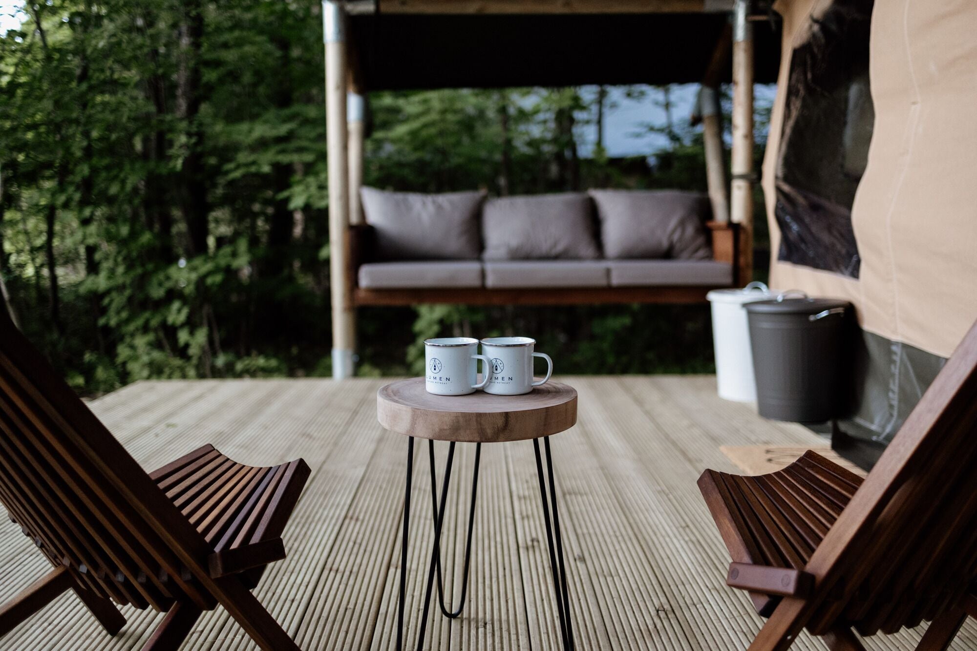 A wooden deck with two lounge chairs, a small round table with two mugs, a hanging swing chair, and a trash bin.