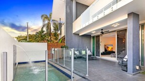 Un área de piscina moderna al aire libre con barandilla de vidrio, zona de estar con sofá y sillones, y vista del atardecer a través de los ventanales del edificio.