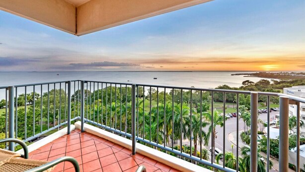 A balcony with a view of the sea at sunset, surrounded by greenery and a paved area with cars. - Coastal Splendor: Seaview ~ Pool ~ Balcony Dining (Darwin City)