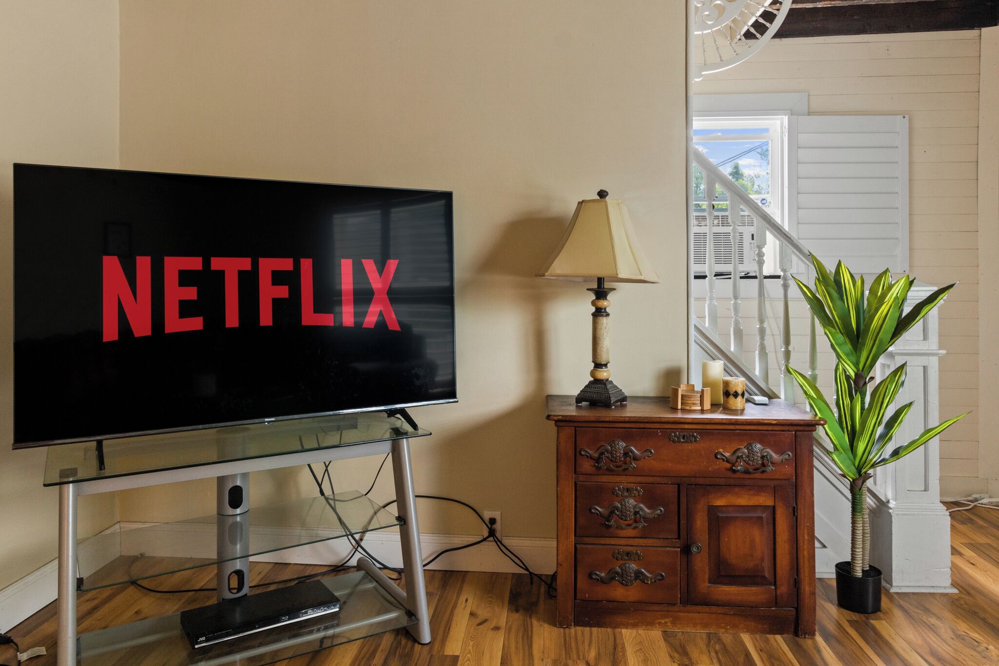A television on a glass stand with the word 'NETFLIX' on the screen, a wooden cabinet with drawers, a lamp, and a potted plant.