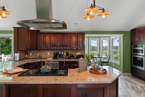 A modern kitchen with dark wood cabinets, a large island with a kettle, and a view of the outdoors through large windows. - Salmon River View! - Sleeps 12 - Bar, Grill and Perfect Location (Pulaski)