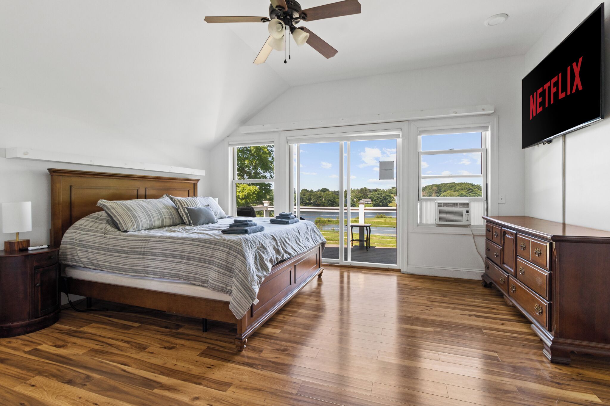 A bedroom with a bed, wooden floor, ceiling fan, TV, and a view of a lake.