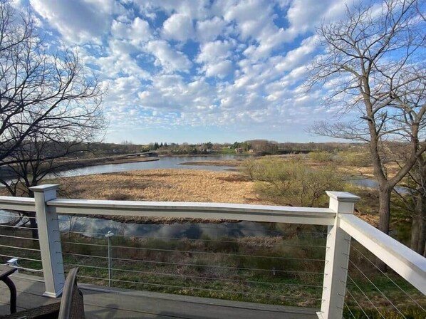 A riverside view from a deck with a railing, a clear sky, and a distant landscape including trees and buildings. - Salmon River View! - Sleeps 12 - Bar, Grill and Perfect Location (Pulaski)