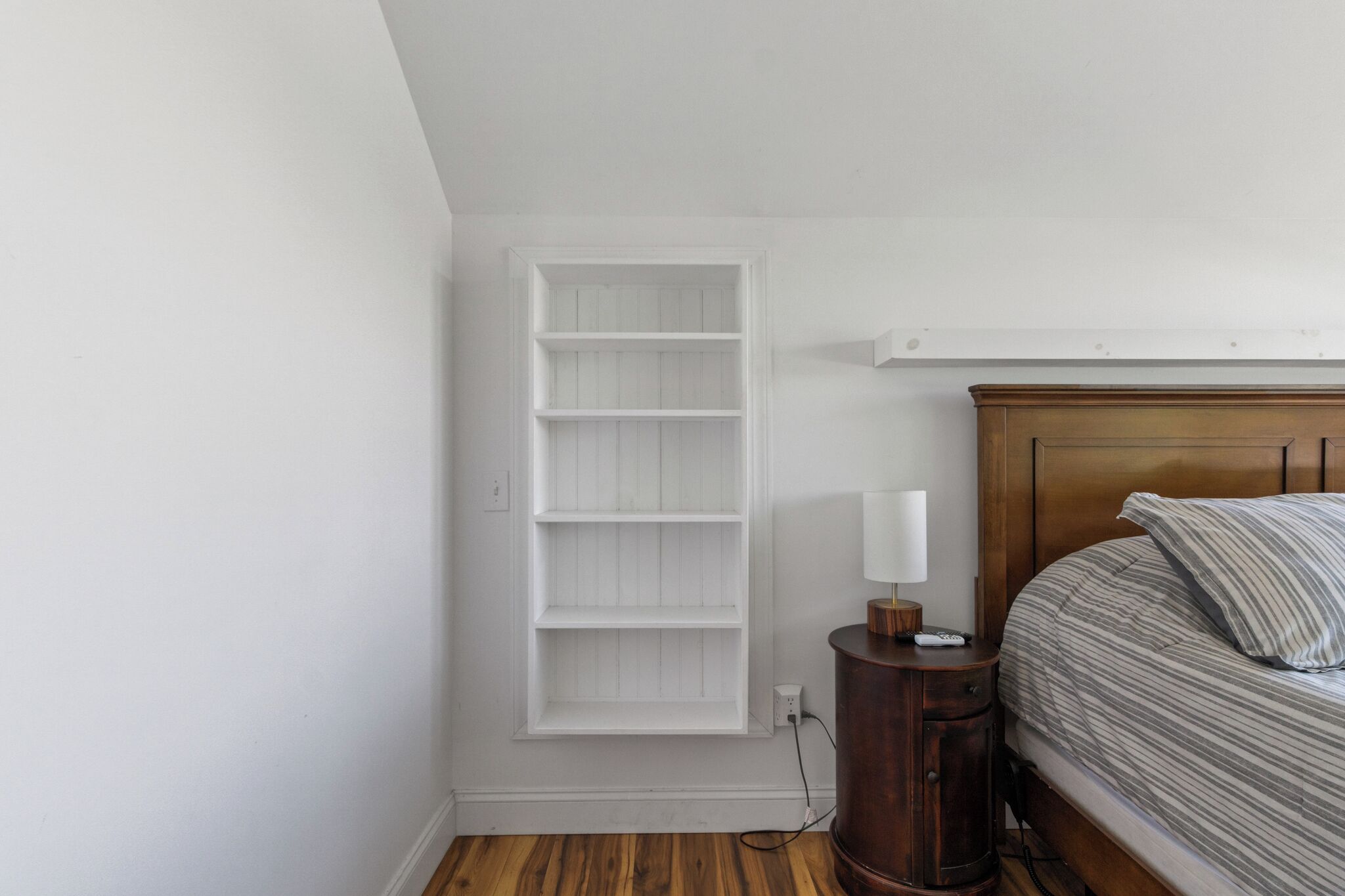 A bedroom with a bed, a nightstand, a wall-mounted shelf, and a wooden floor.