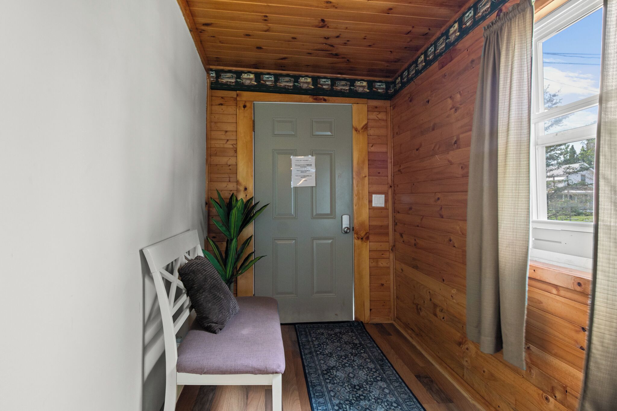 A wooden interior hallway with a door, a bench with a cushion, a potted plant, and a rug.