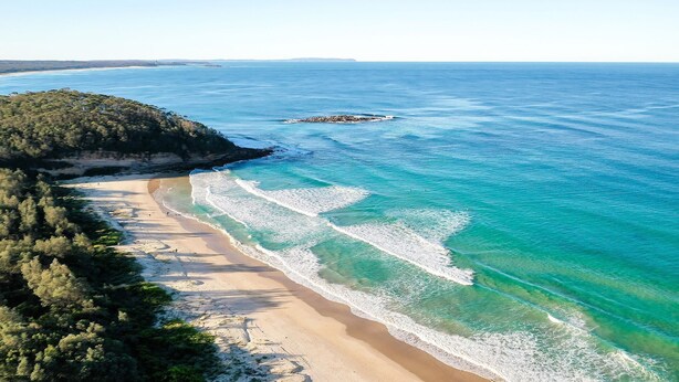 Un paisaje costero con una playa arenosa, aguas turquesas cristalinas y un promontorio rocoso a lo lejos.