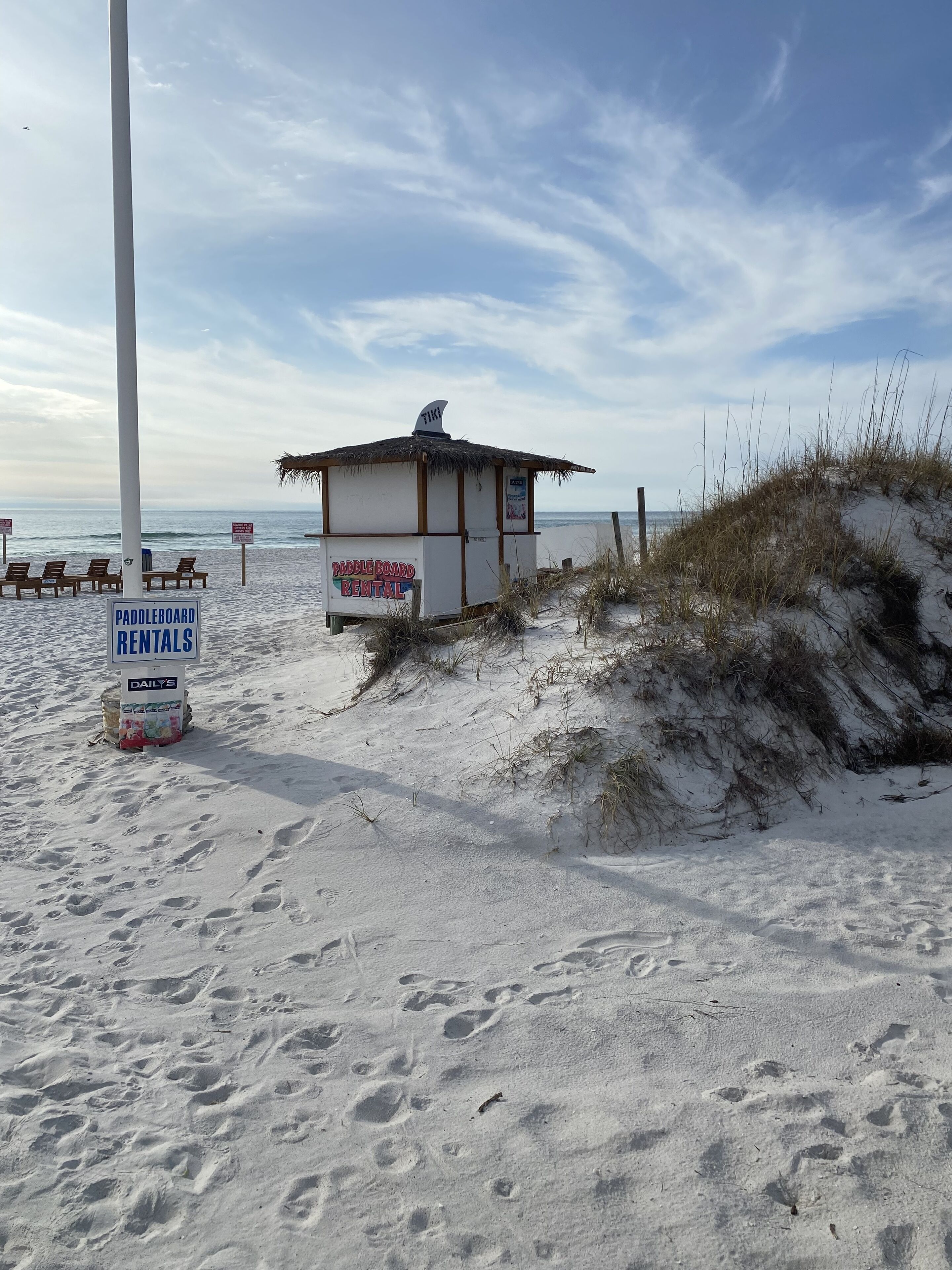 Una cabaña de alquiler frente a la playa con techo de paja, un cartel de alquiler de tablas de remo y una zona con sillas de playa.