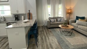 A modern kitchen with white cabinets, a marble countertop, and a coffee maker. There is a dining area with blue chairs and a gray rug. A gray sofa and a coffee table are in the living room. - Family Friendly Venture Out Getaway  (Panama City Beach)