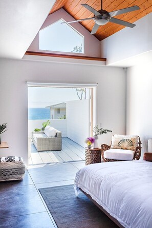 Une chambre avec un grand lit, un ventilateur de plafond et une vue sur la mer, visible par une porte ouverte.