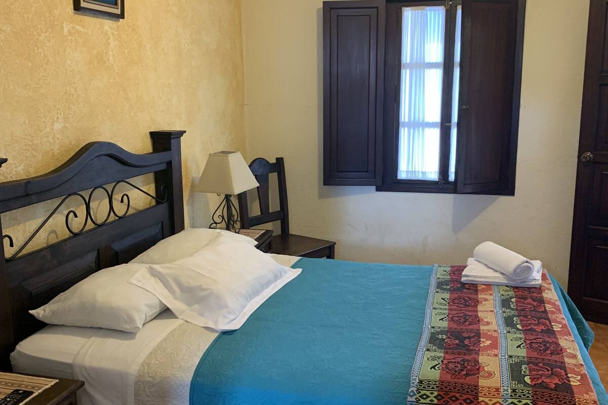 Quarto com cama, mesa de cabeceira, luminária, janela com cortinas e um quadro na parede.