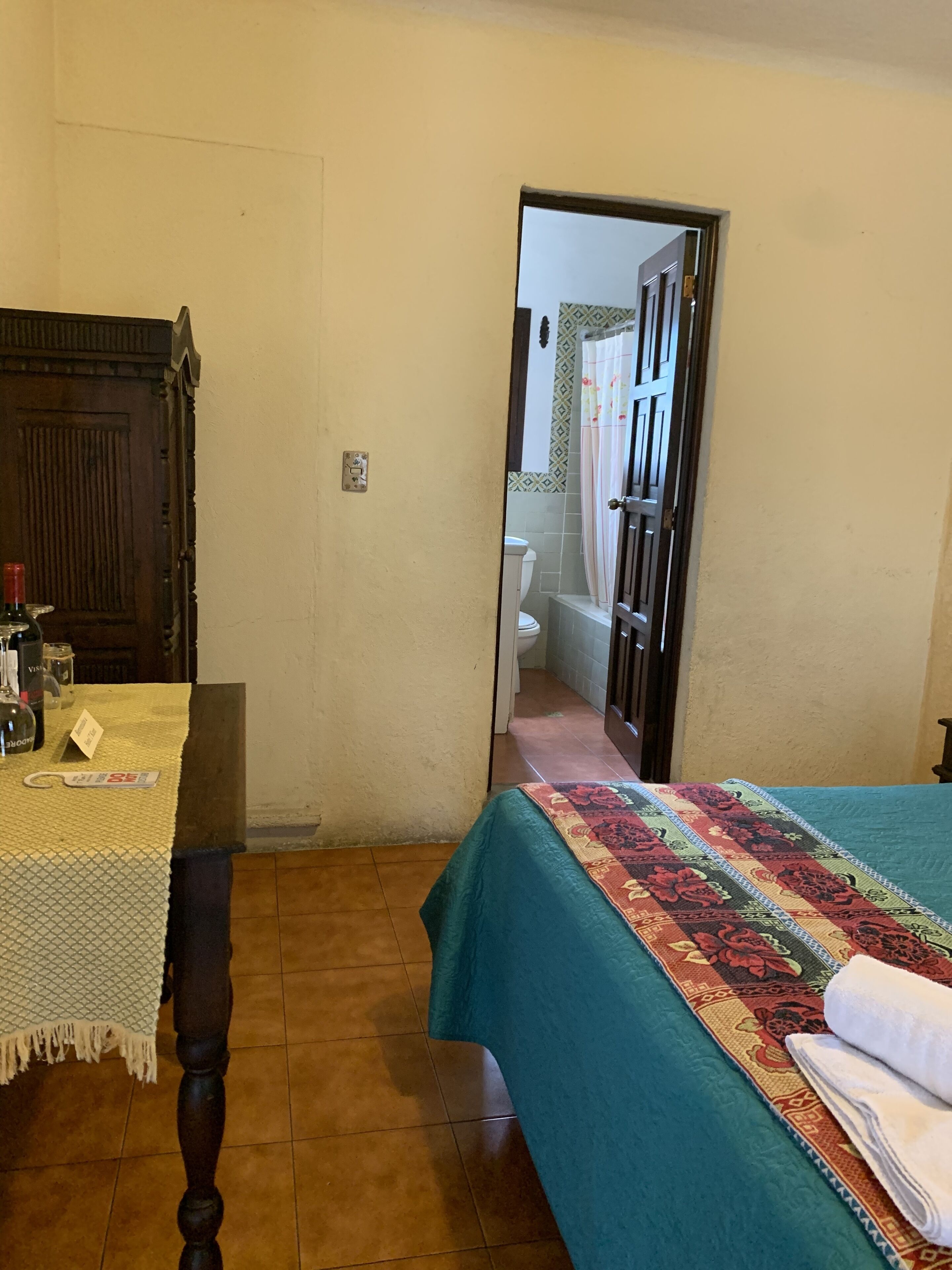 Une pièce avec une table en bois, un lit recouvert d’une courtepointe à motifs et une porte donnant sur une salle de bain.